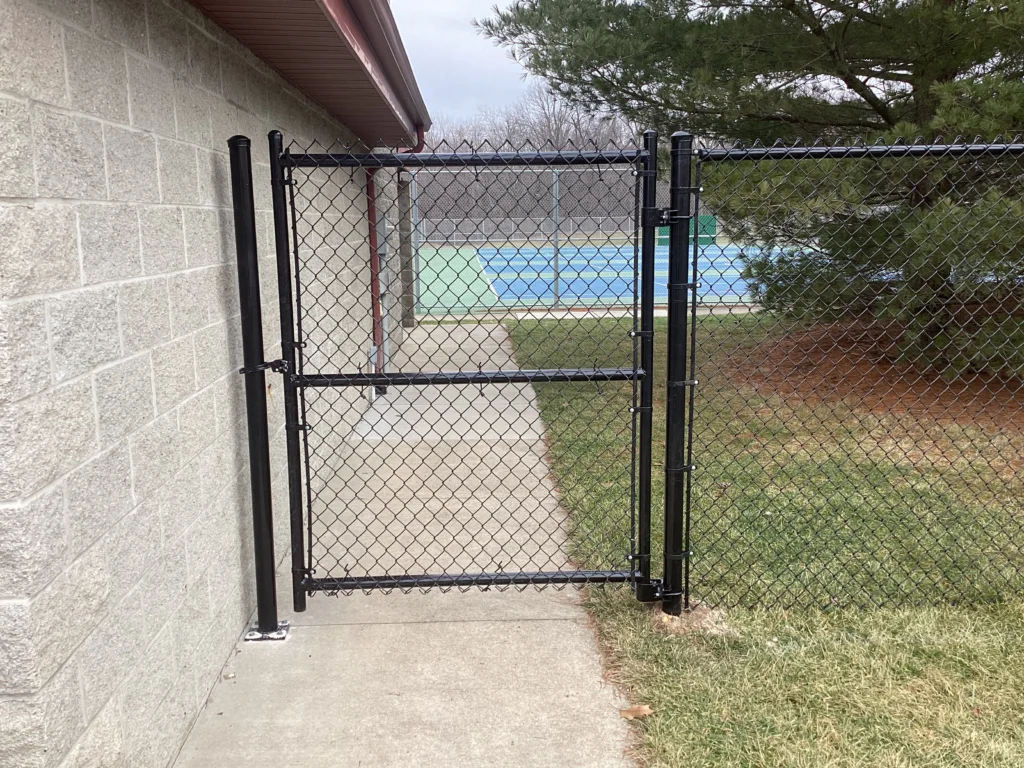 A 6-foot tall black vinyl-coated chain-link gate and fence bordering a concrete walkway next to a building