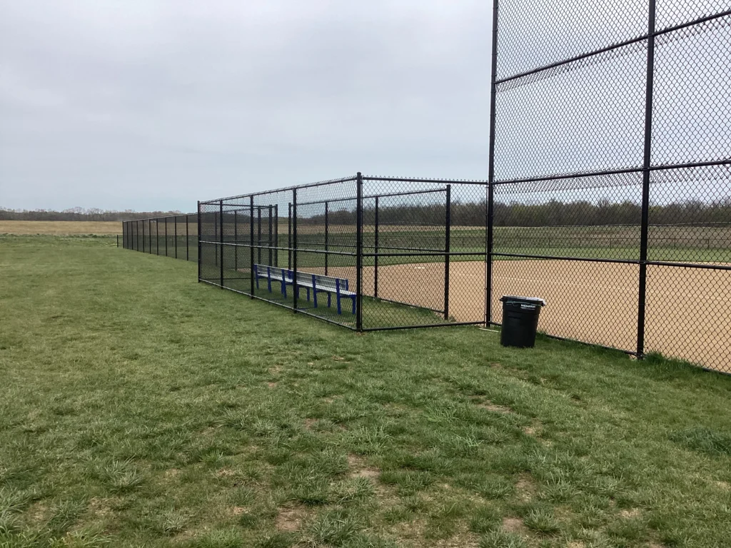 A baseball field dugout area enclosed with black vinyl-coated chain-link fencing, including a bench and nearby trash receptacle
