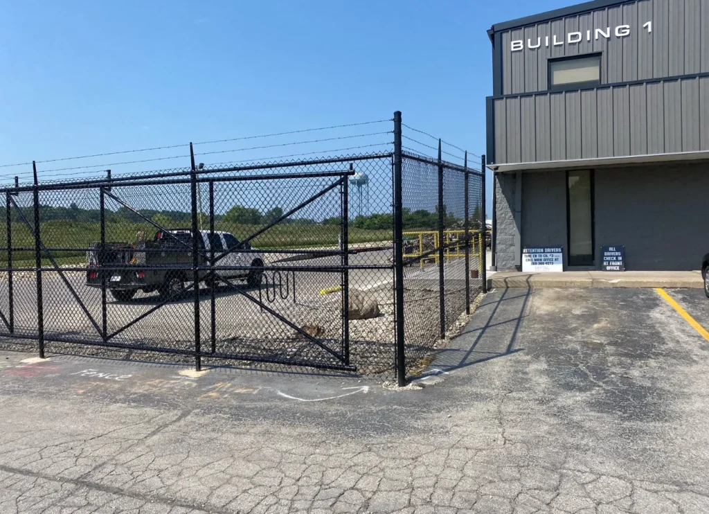 A black vinyl-coated chain-link security fence with a cantilever slide gate and barbed-wire topper protecting the parking area of an industrial building