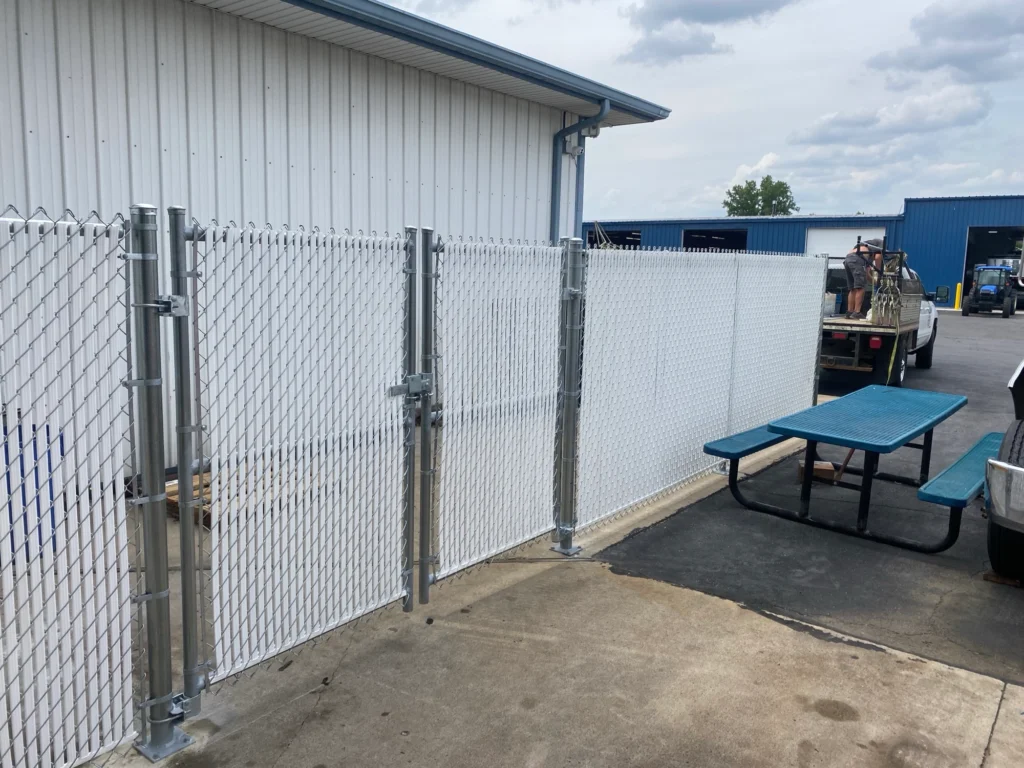 A commercial chain-link fence with white privacy slats and a double swing gate enclosing a concrete area next to a white industrial building