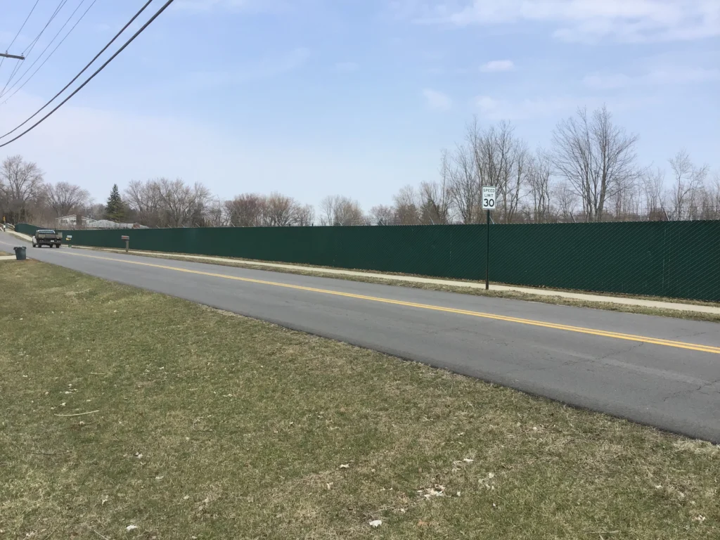 A continuous perimeter security fence featuring dark green privacy slats installed along a two-lane paved road