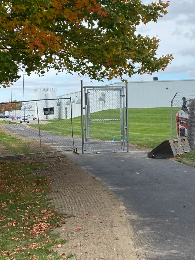 A galvanized chain-link pedestrian gate with a security frame installed across an asphalt pathway leading to a large industrial building