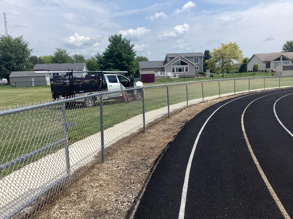 A galvanized chain-link perimeter fence running alongside a black asphalt running track with residential houses in the background