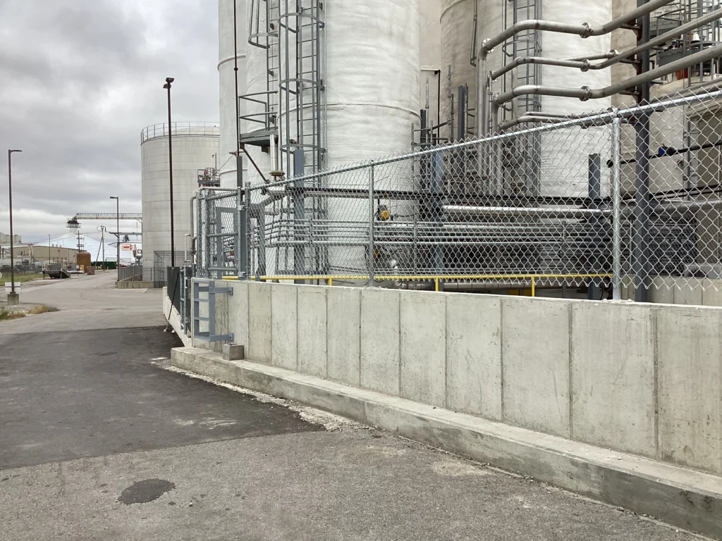 A galvanized chain-link security fence with a top rail installed on top of a concrete retaining wall at an industrial facility with large storage tanks