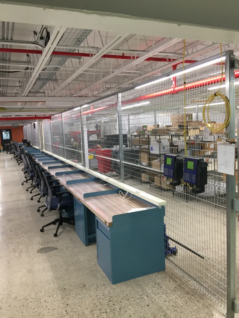 A long industrial partition made of galvanized welded wire mesh separating a row of blue workstations from a warehouse storage area