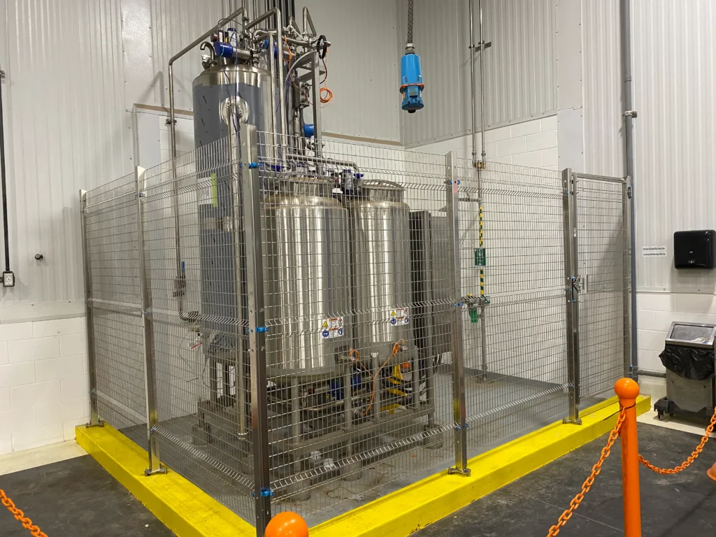 A polished stainless steel wire mesh safety partition with integrated gates enclosing industrial machinery on a yellow-bordered concrete floor