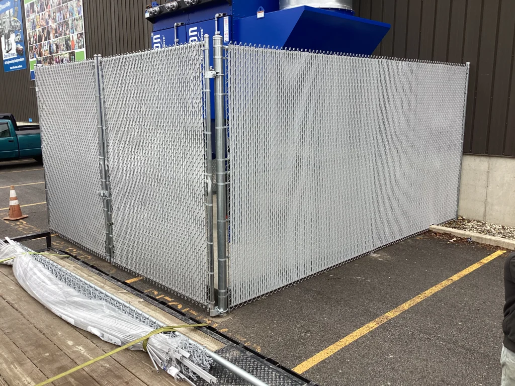 A tall galvanized chain-link security enclosure with gray privacy slats protecting outdoor industrial equipment